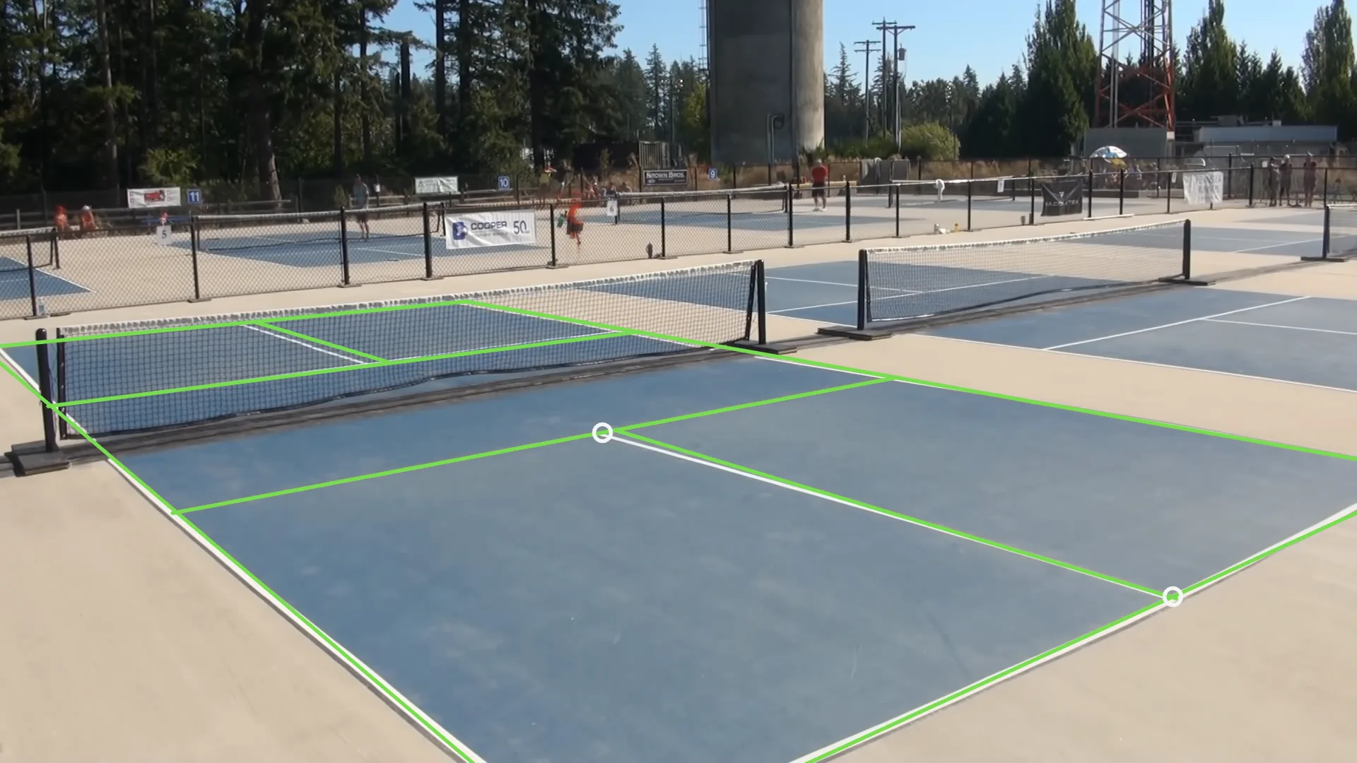 An outdoor pickleball court seen from beside and behind the near baseline, with the full court model drawn in green over the painted lines, and two circled held-out corners