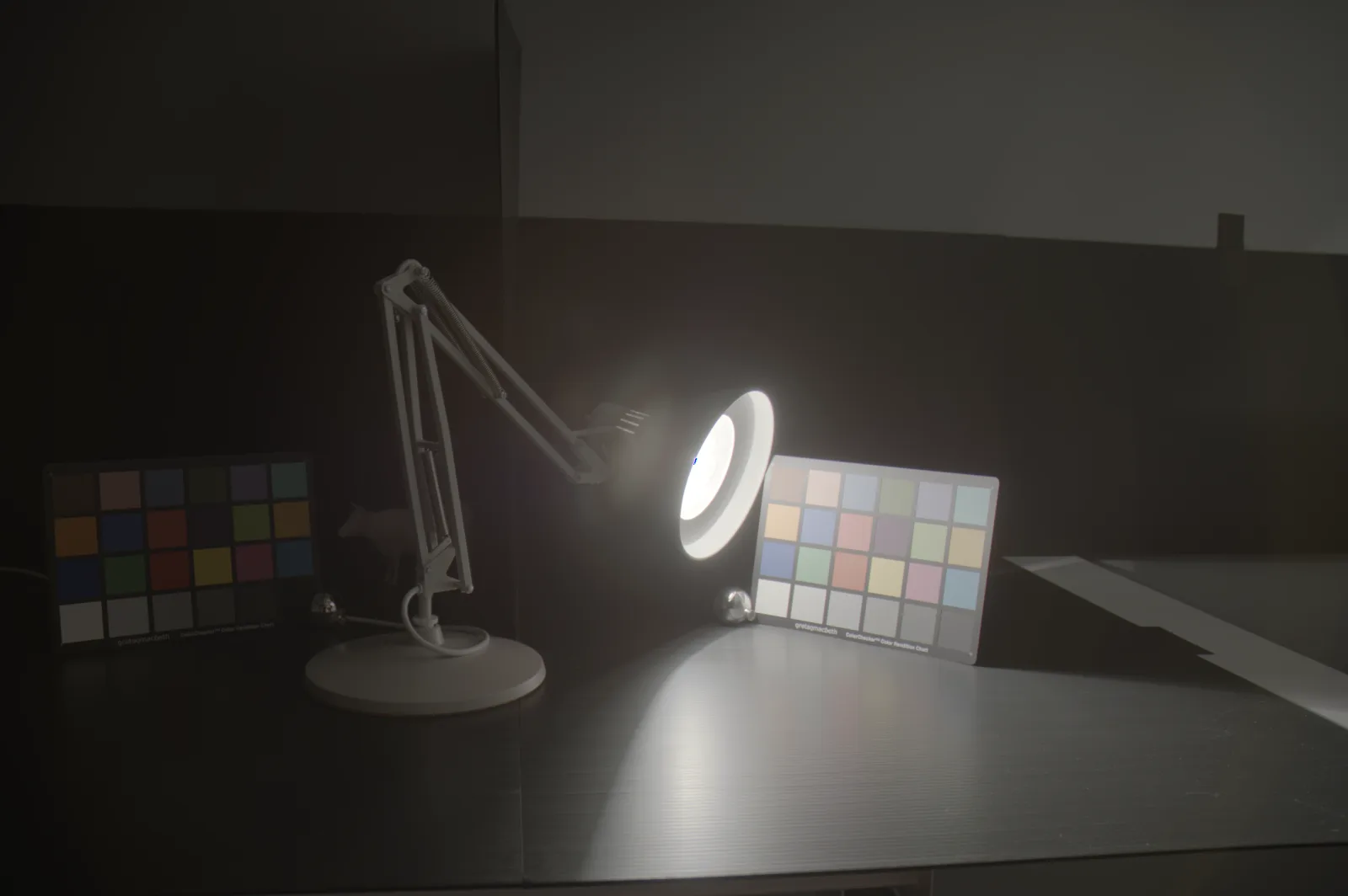 A desk lamp with a lit bulb between two ColorChecker charts, one brightly lit and one in deep shadow, on a dark table